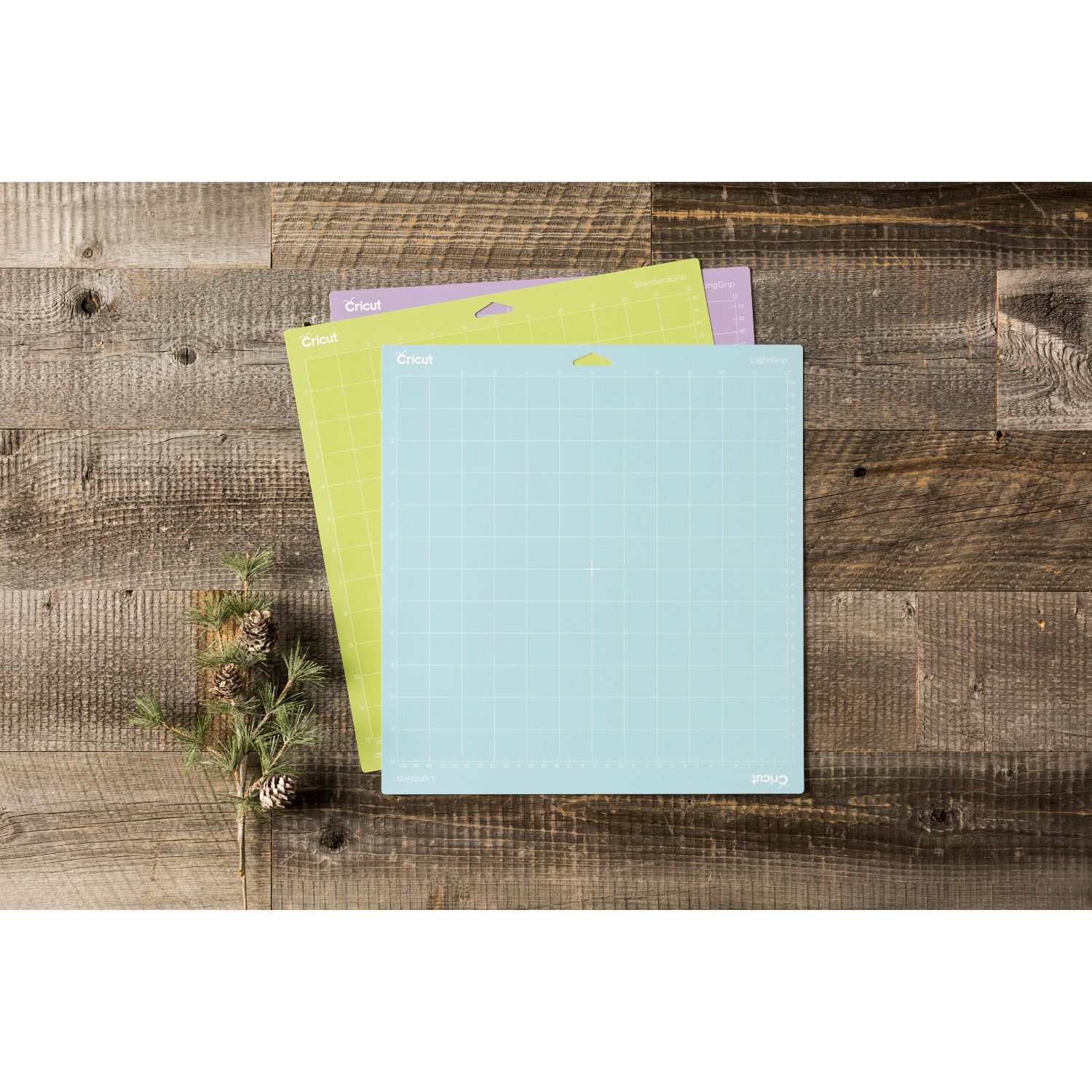 Three Cricut cutting mats with a pine branch and a wooden background.