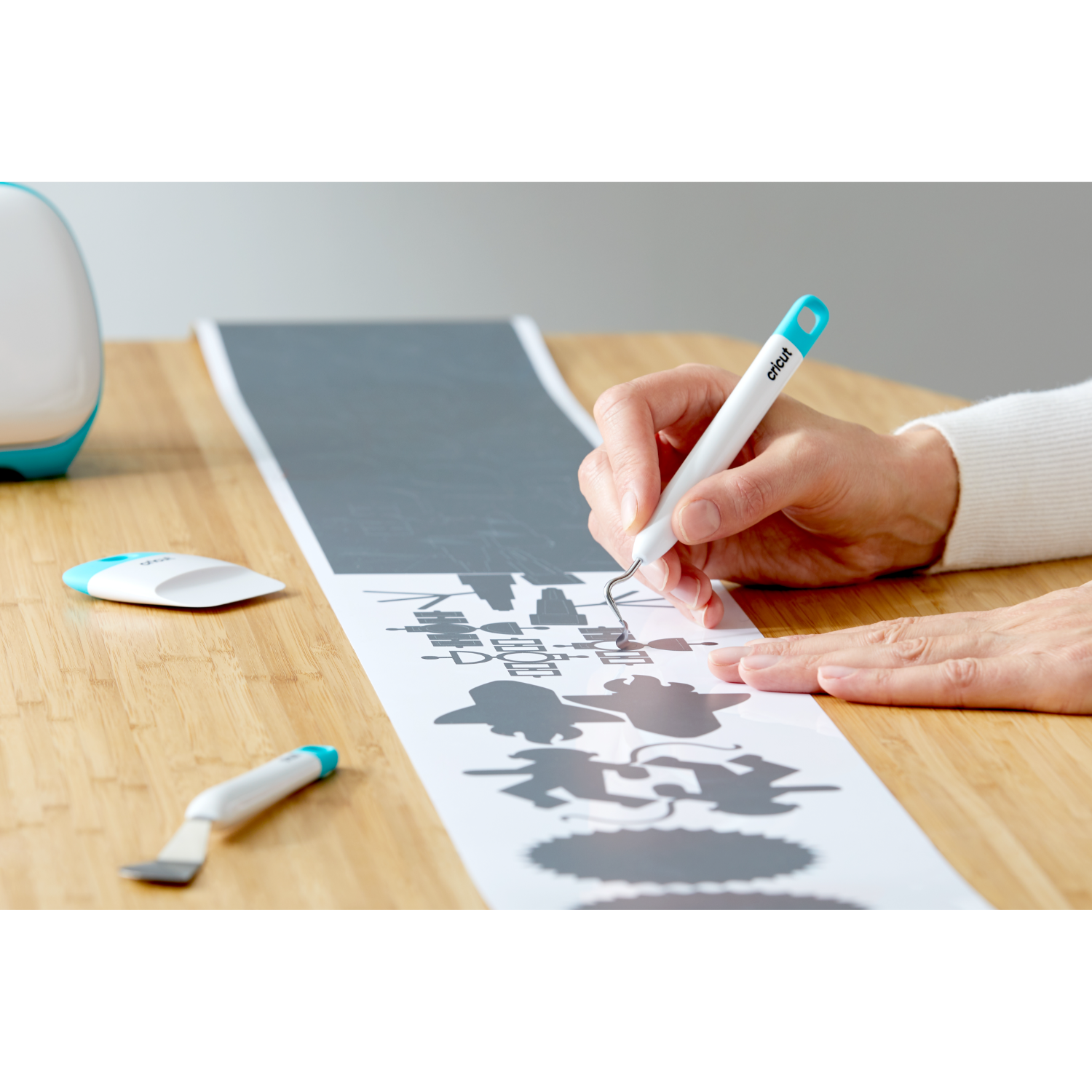 A person's hand using a Cricut tool to weed a gray design from white material on wood.