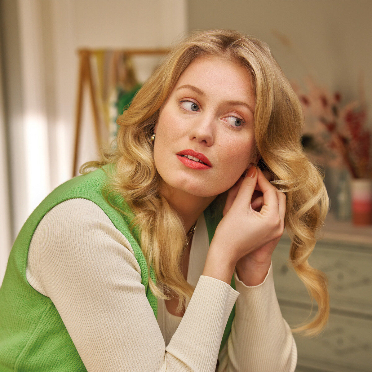 Blondynka zakłada kolczyk, patrząc w prawo. Ma na sobie zieloną kamizelkę.