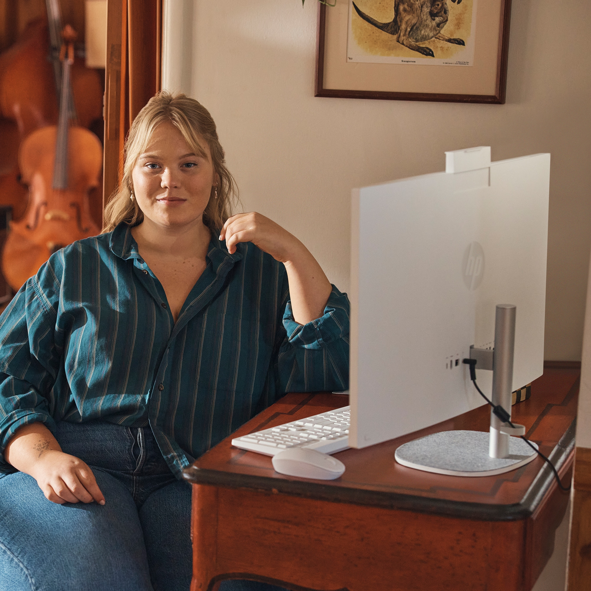Blonde Frau an einem Schreibtisch mit Computer. Hintergrund: Cello, Gemälde und Holztisch.