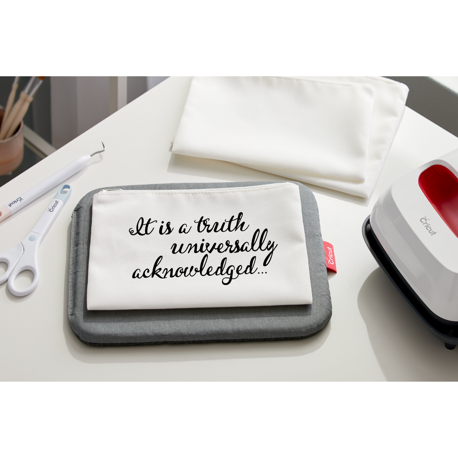 Een witte tafel met Cricut-gereedschap en een tas met de tekst: “It is a truth universally acknowledged...”
