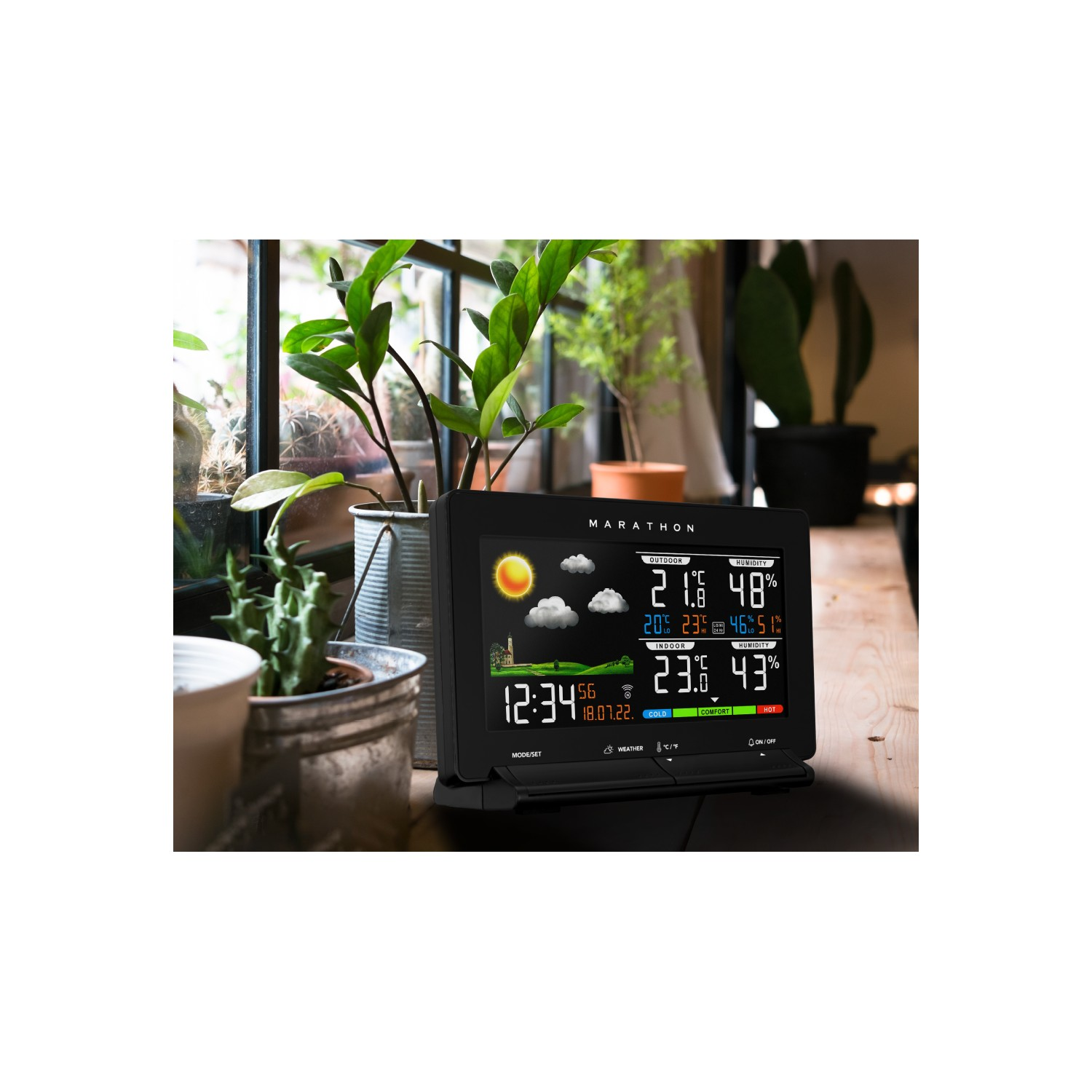 A black weather station displaying weather data, with a sunny icon, on a wooden table near plants.