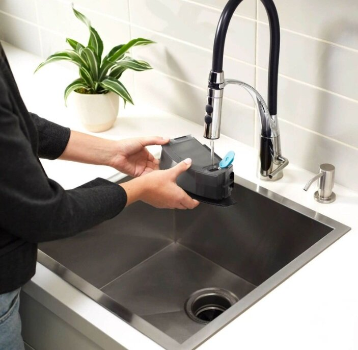 Osoba trzymająca czarny pojemnik pod kranem w zlewie ze stali nierdzewnej.