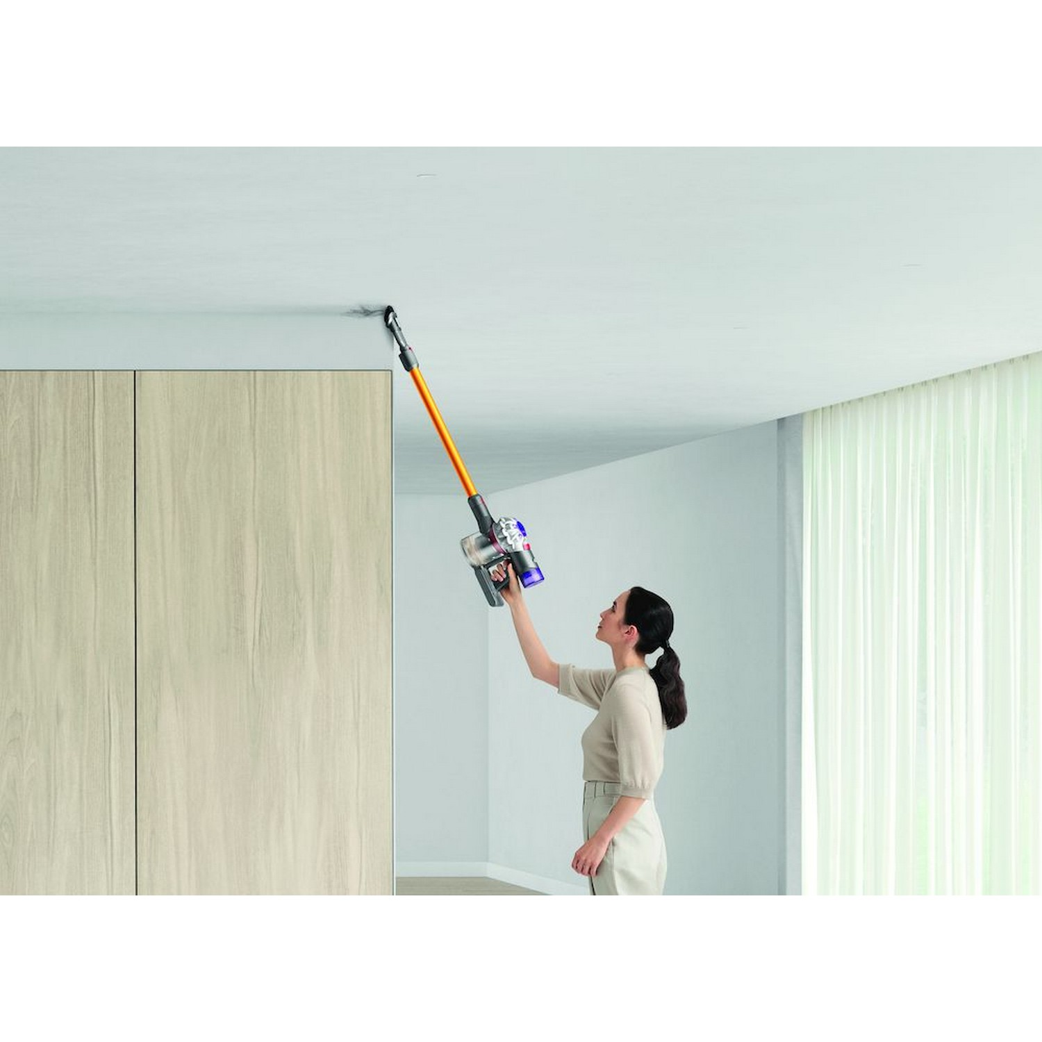 Een vrouw die een plafond stofzuigt met een draadloze stofzuiger. Een houten kast staat links.