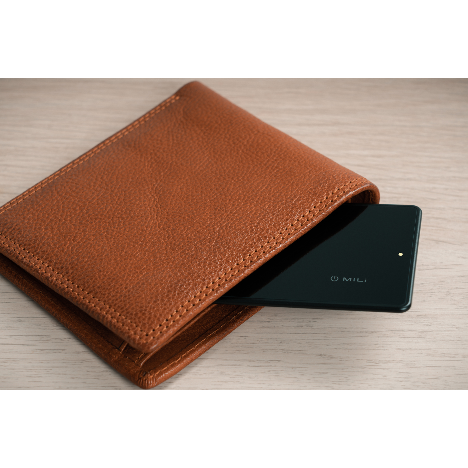 A brown leather wallet with a black tablet partially inserted, resting on a light wooden surface.