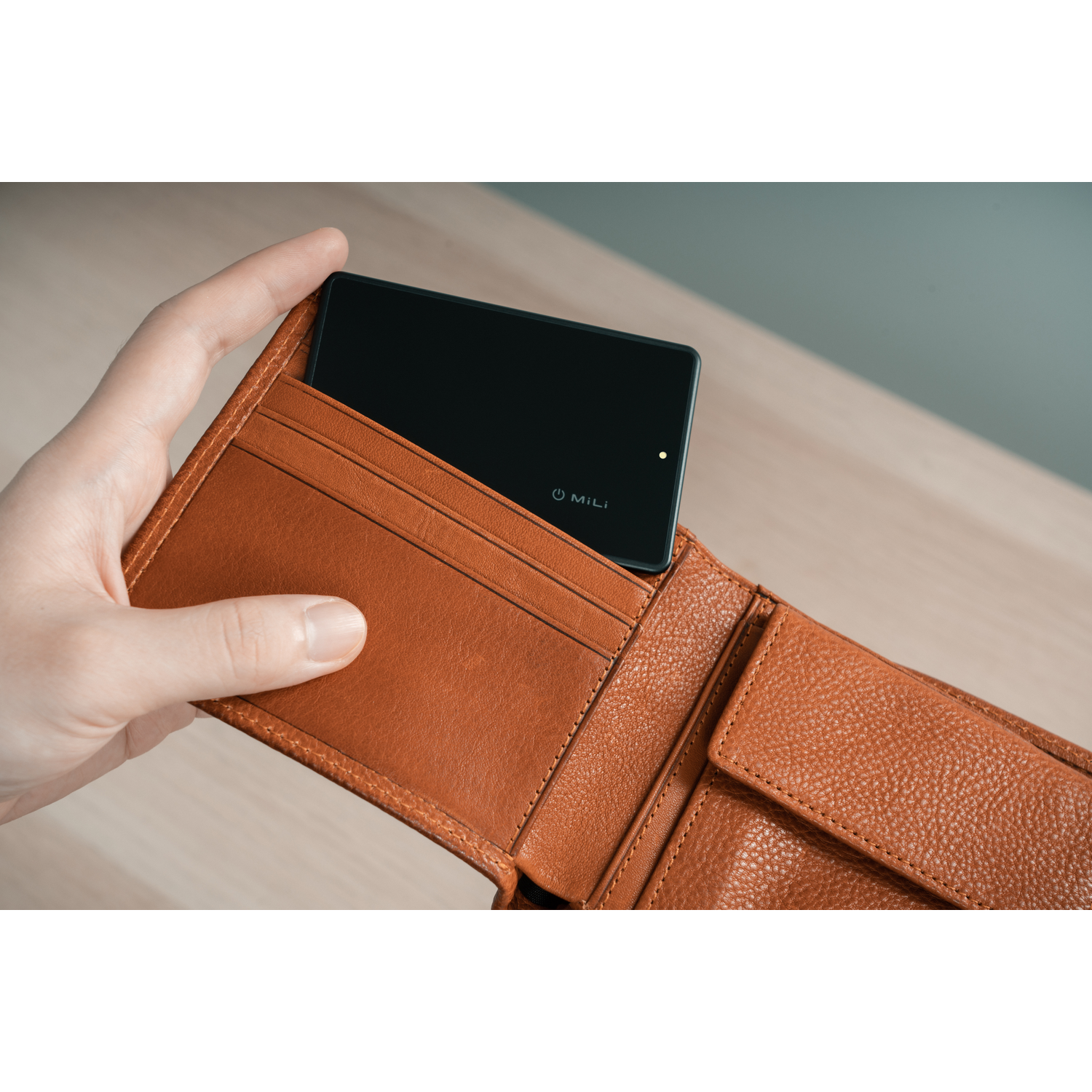 Hand putting a black phone into a brown leather wallet with the word MILI on phone.