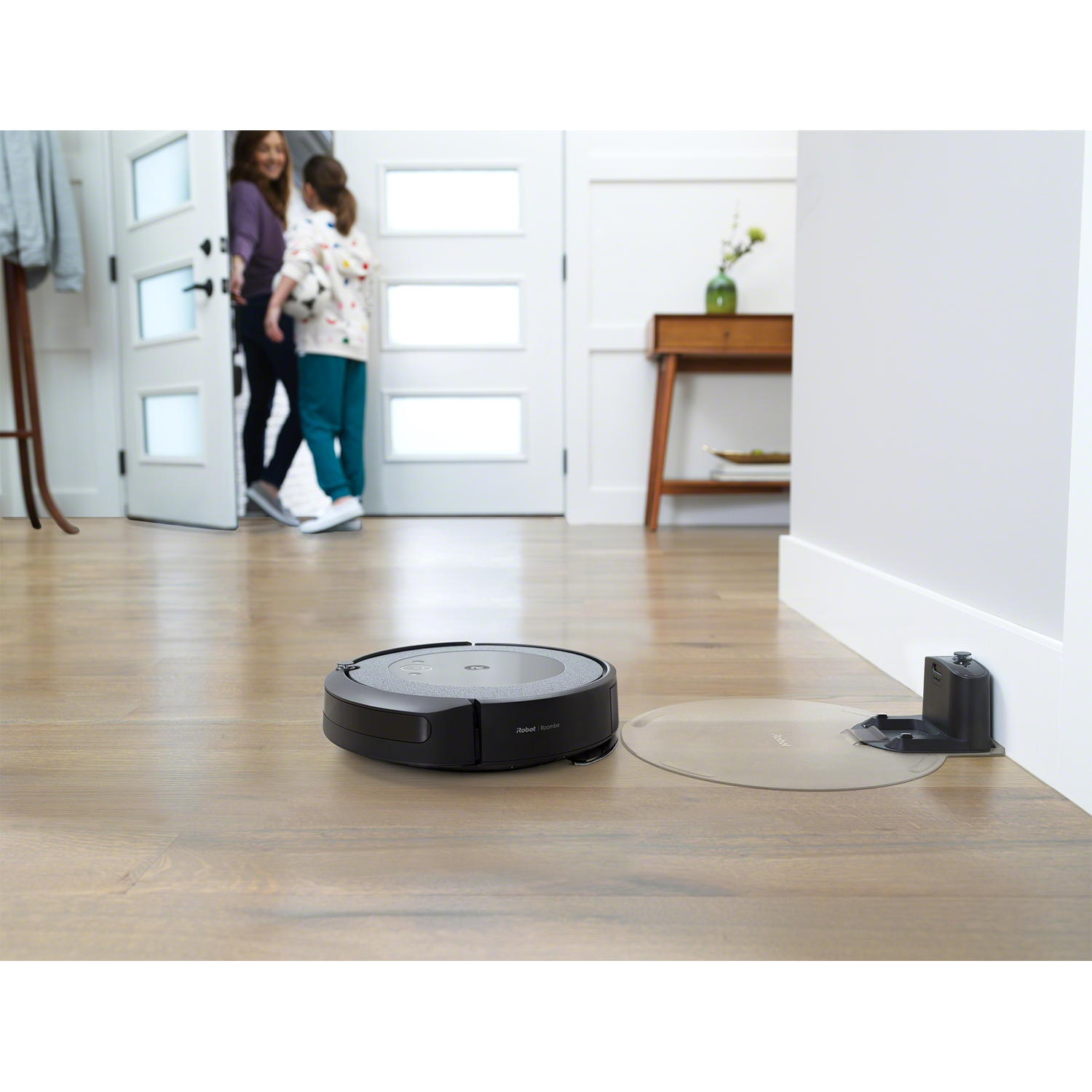 Robotstofzuiger op houten vloer. Twee vrouwen komen de kamer binnen.