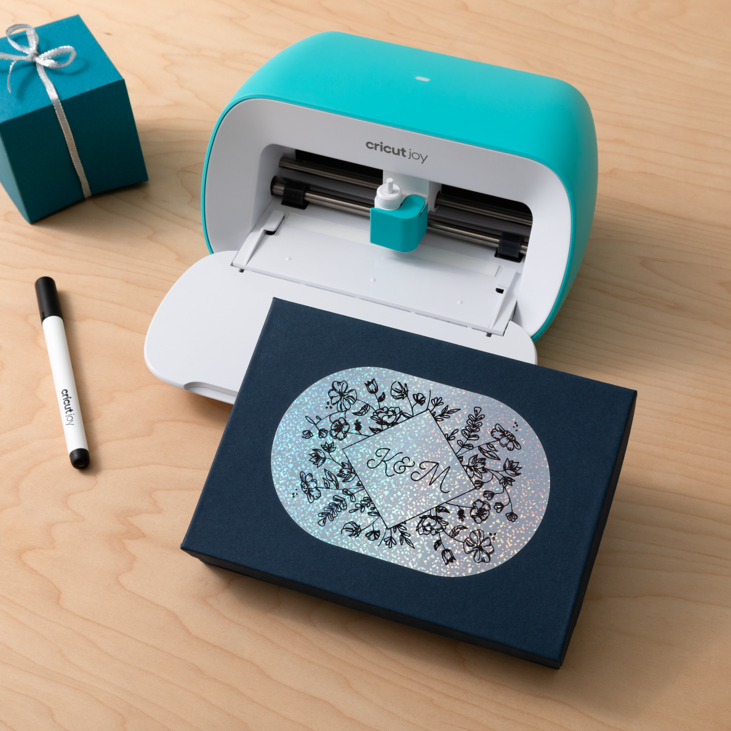 A teal Cricut Joy machine. A box is under. A pen beside. A box with a printed design.