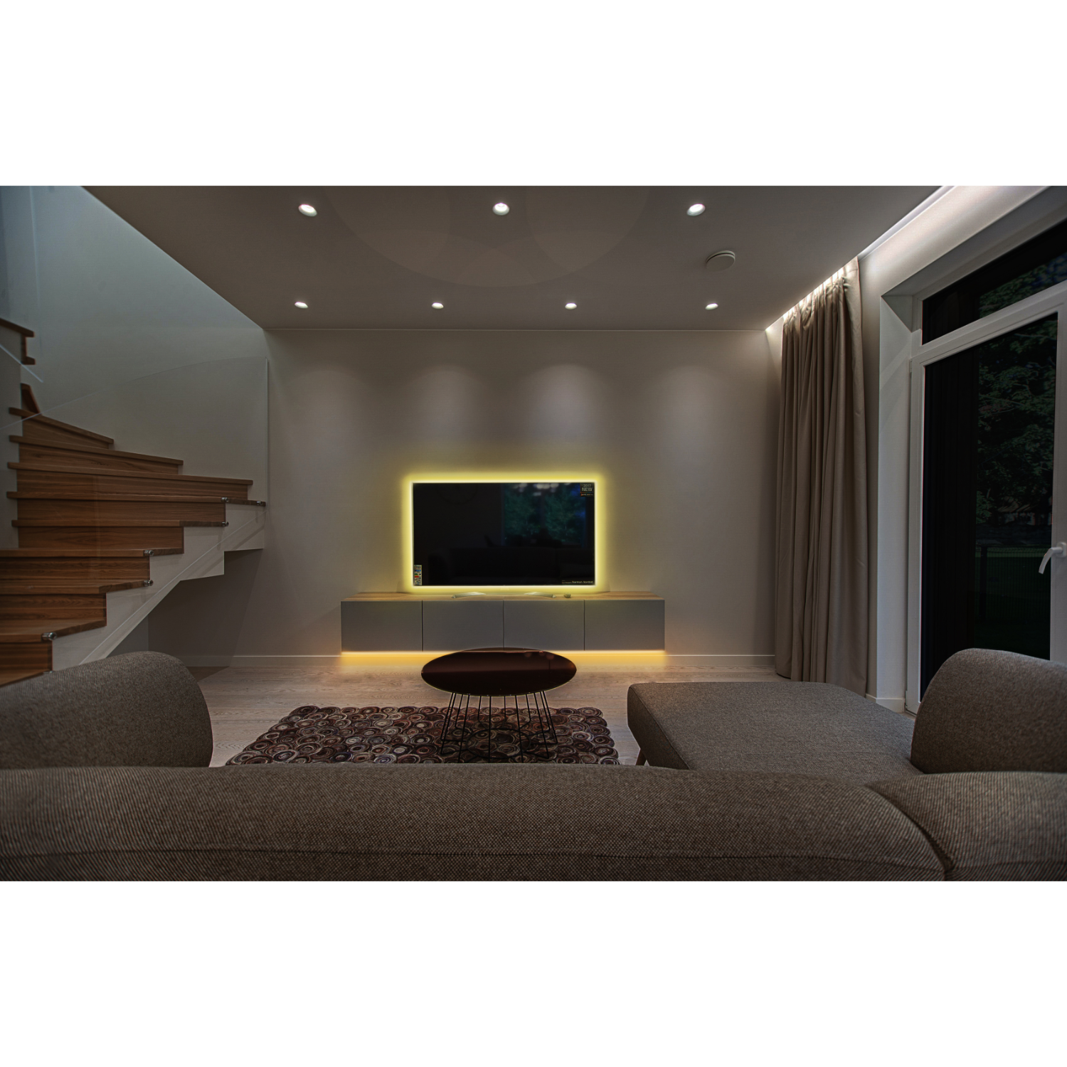 Living room with sofa, TV, and stairs. The TV is mounted on a cabinet, and there is a dark rug.