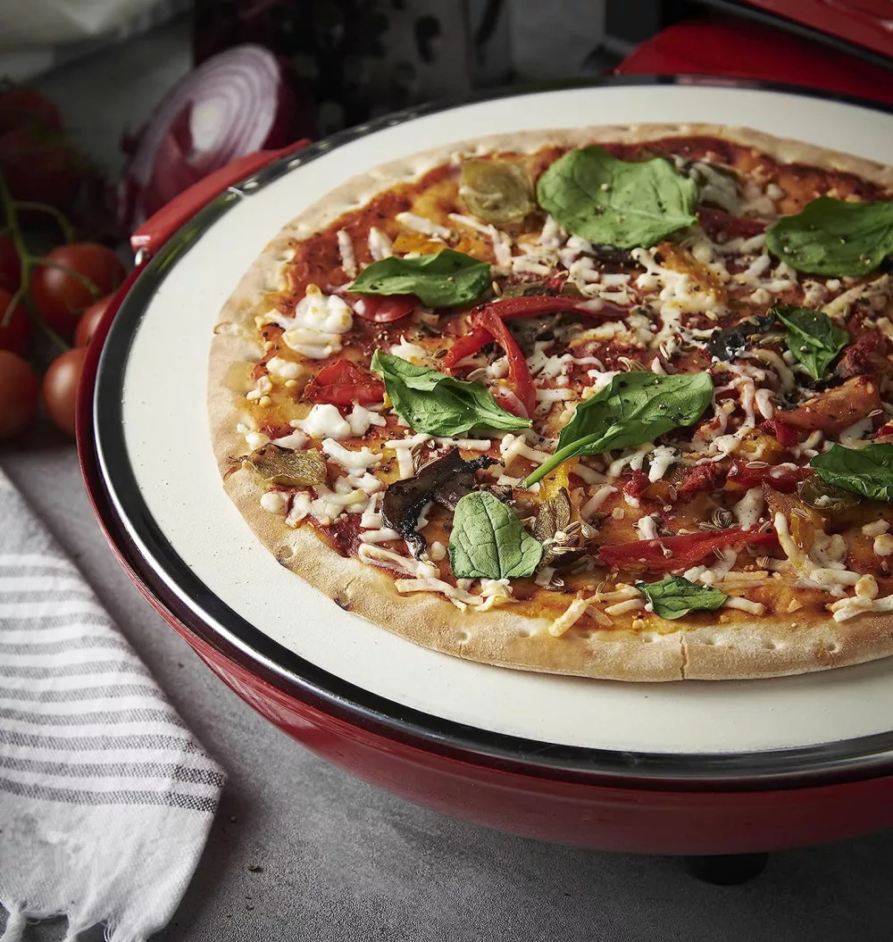 Pizza feltétekkel egy fehér kövön egy piros pizzasütőben, szürke felületen.
