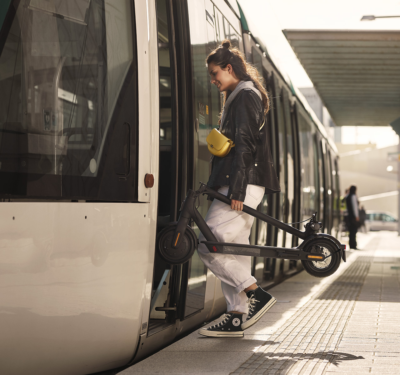 Frau verlässt eine Straßenbahn, hält einen zusammengeklappten schwarzen Roller. Sie trägt eine schwarze Jacke und weiße Hose.