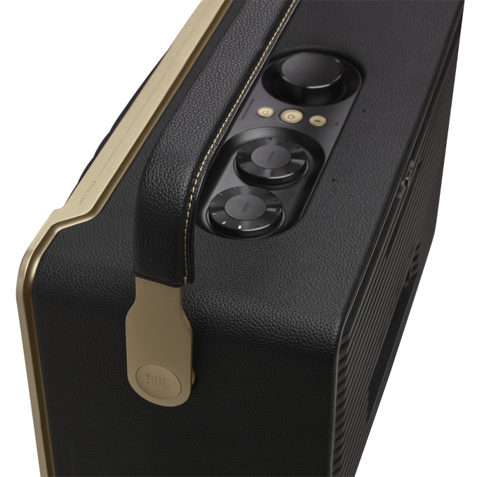 Altavoz JBL negro y dorado. Tiene controles de volumen, graves y agudos. Asa de cuero y ribete dorado.