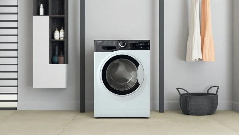 Biała pralka stoi w łazience, z koszem i wiszącymi ubraniami w pobliżu. Szafka jest widoczna po lewej stronie.