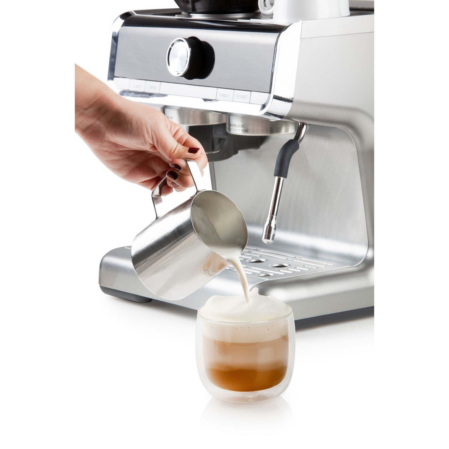 A person pours milk foam into a glass of coffee from a silver pitcher. A machine is in the background.