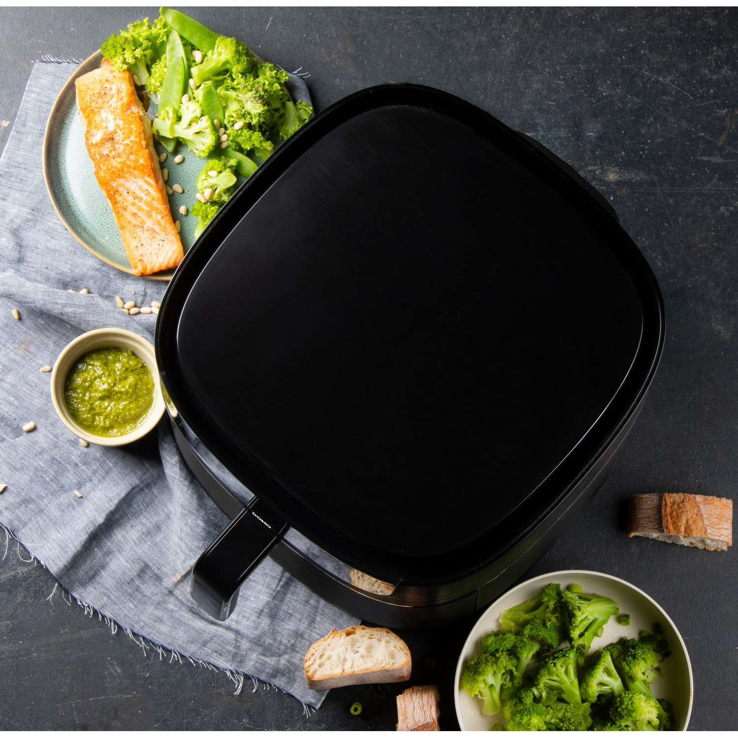 Friteuse à air noire avec de la nourriture, dont du saumon, du brocoli et du pain sur une table.