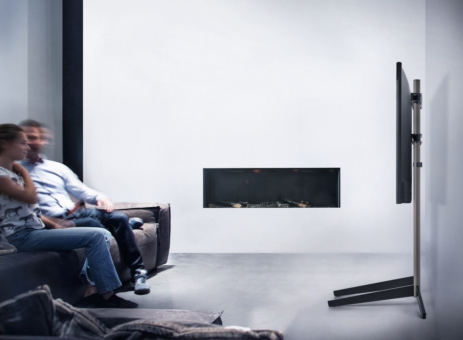 Ein Paar sitzt auf einem Sofa und sieht fern. Ein großer Bildschirm-TV steht auf einem Ständer. Im Hintergrund befindet sich ein Kamin.