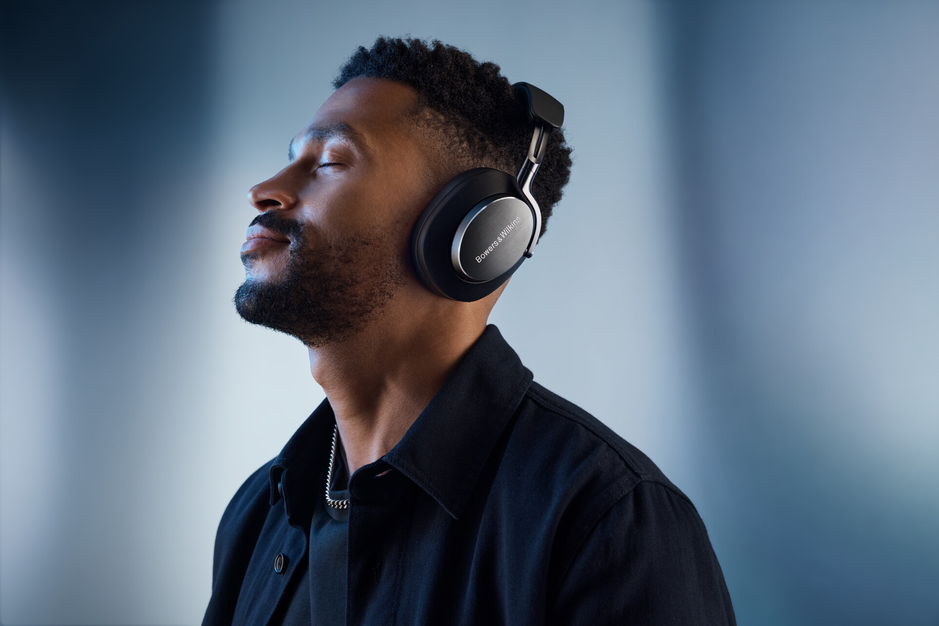 Mann mit geschlossenen Augen, der schwarze Kopfhörer und ein schwarzes Hemd trägt. Blauer Hintergrund.