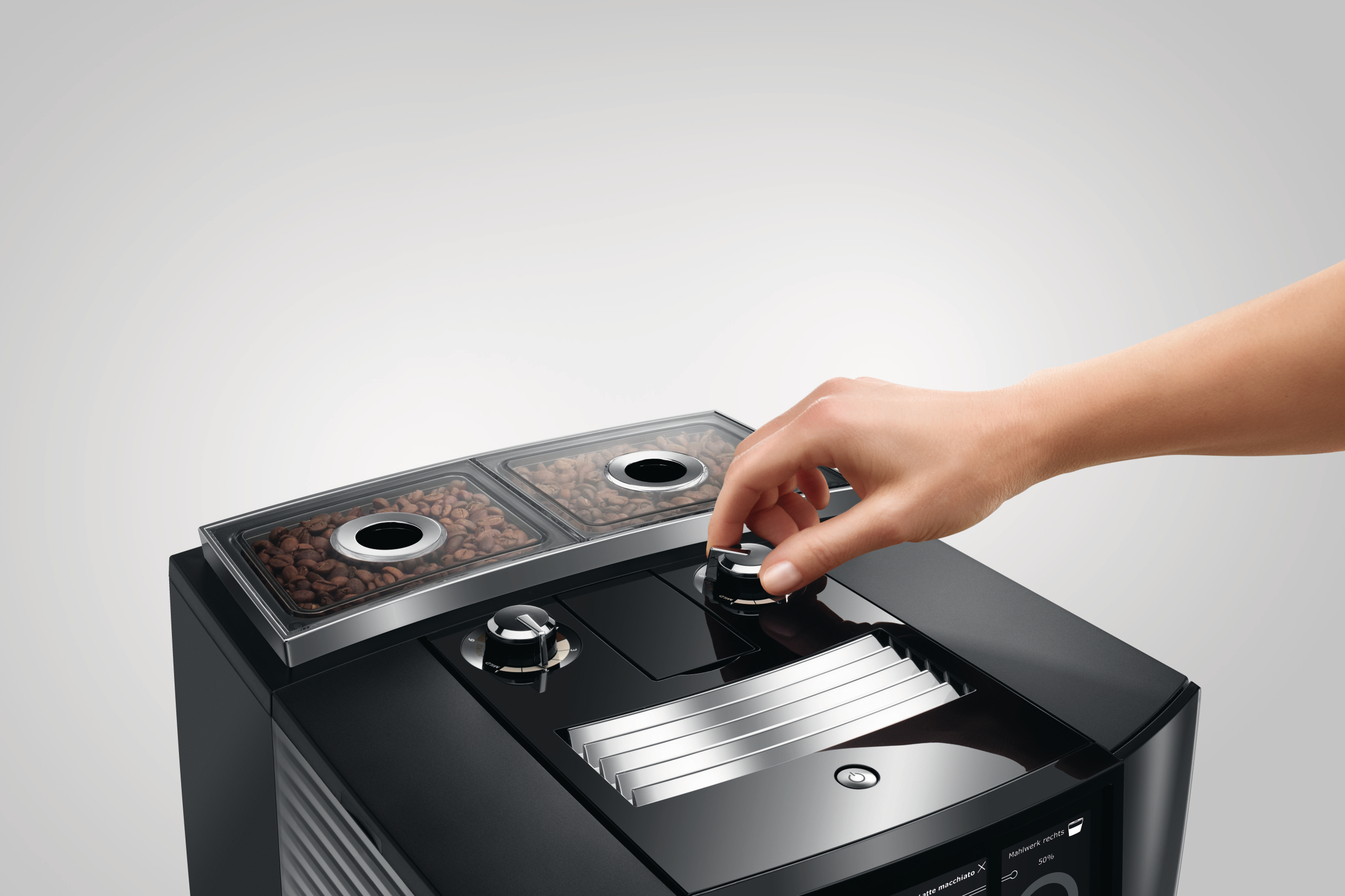 Hand dreht einen Drehregler an einer schwarzen Kaffeemaschine. Zwei Bohnenbehälter oben.