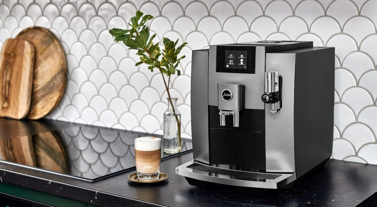 Ekspres do kawy na blacie z latte przed nim. Biała ściana z kafelkami, drewniana deska do krojenia.