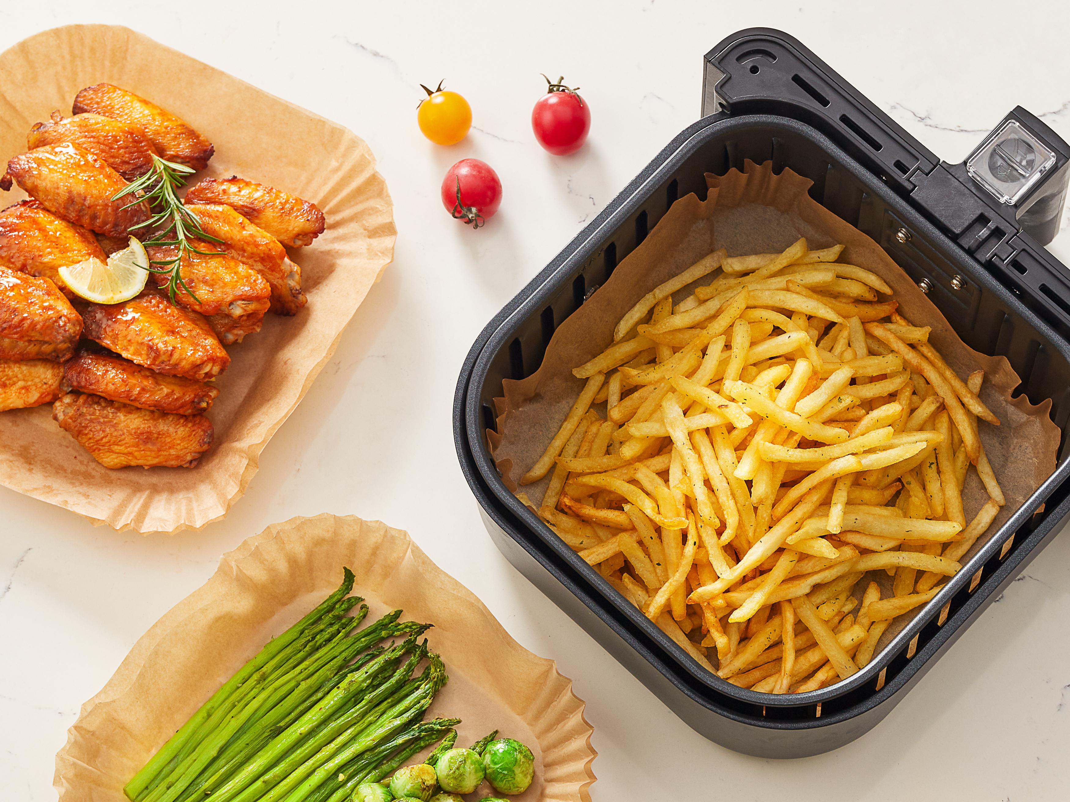 Eine Heißluftfritteuse mit Hähnchenflügeln, Pommes Frites, Spargel und Tomaten auf Backpapier.