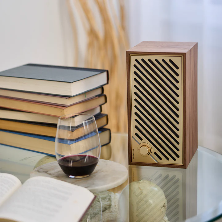 Une enceinte en bois, un verre de vin rouge et une pile de livres sont posés sur une table. Image réfléchie sur la table. Fond beige.