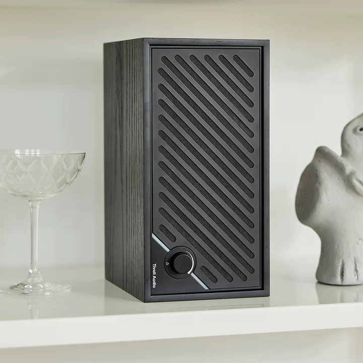 Un haut-parleur noir avec une grille à motifs diagonaux est posé sur une étagère. Une coupe de champagne et une statue de chaque côté.