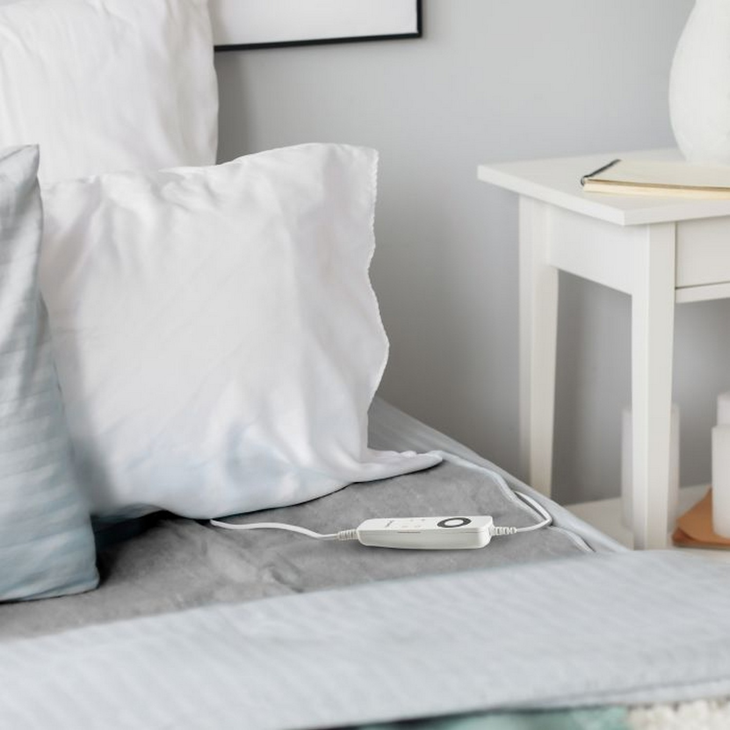 Lit avec oreillers, commande de coussin chauffant, table de chevet blanche. Literie grise et blanche.