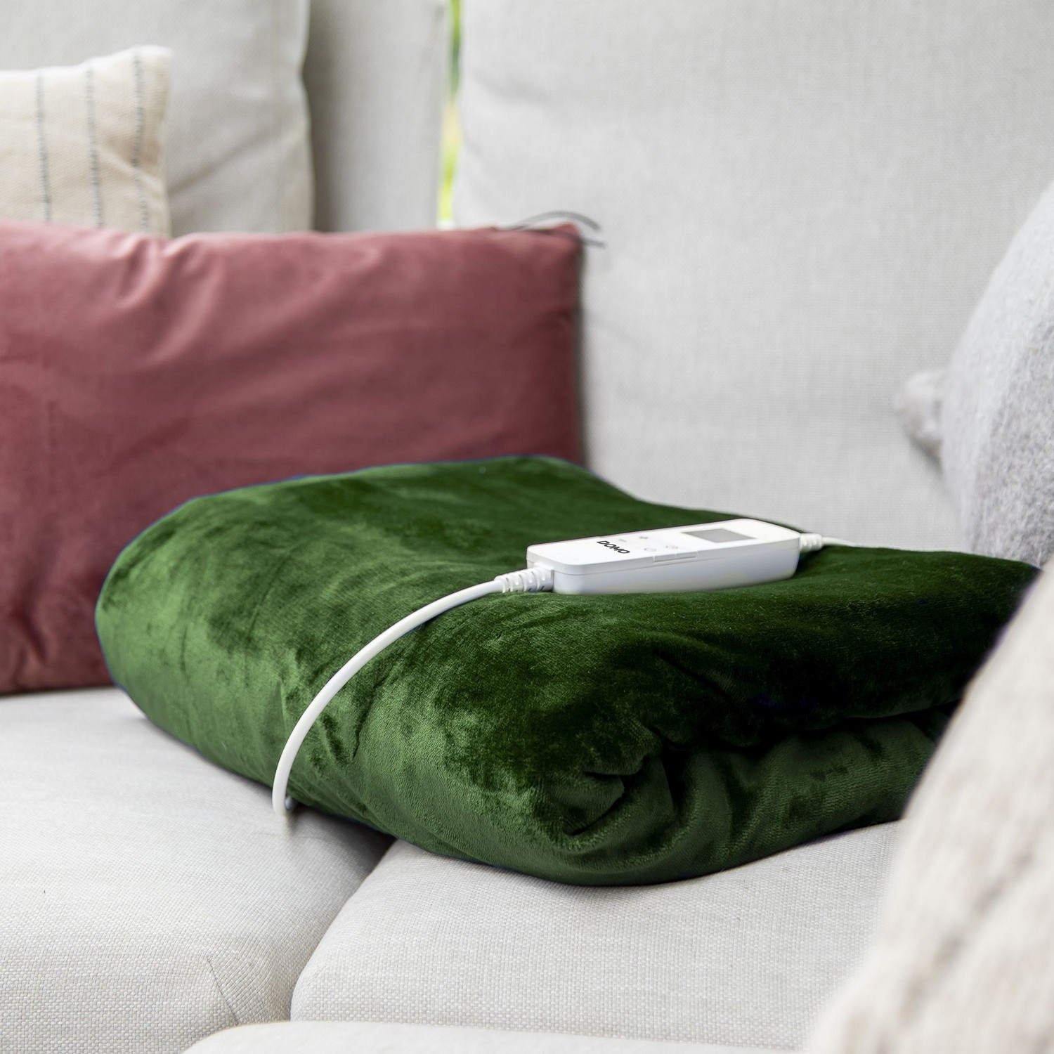 Couverture chauffante verte sur un canapé. Cordon blanc avec panneau de commande. Coussins roses et blancs visibles.