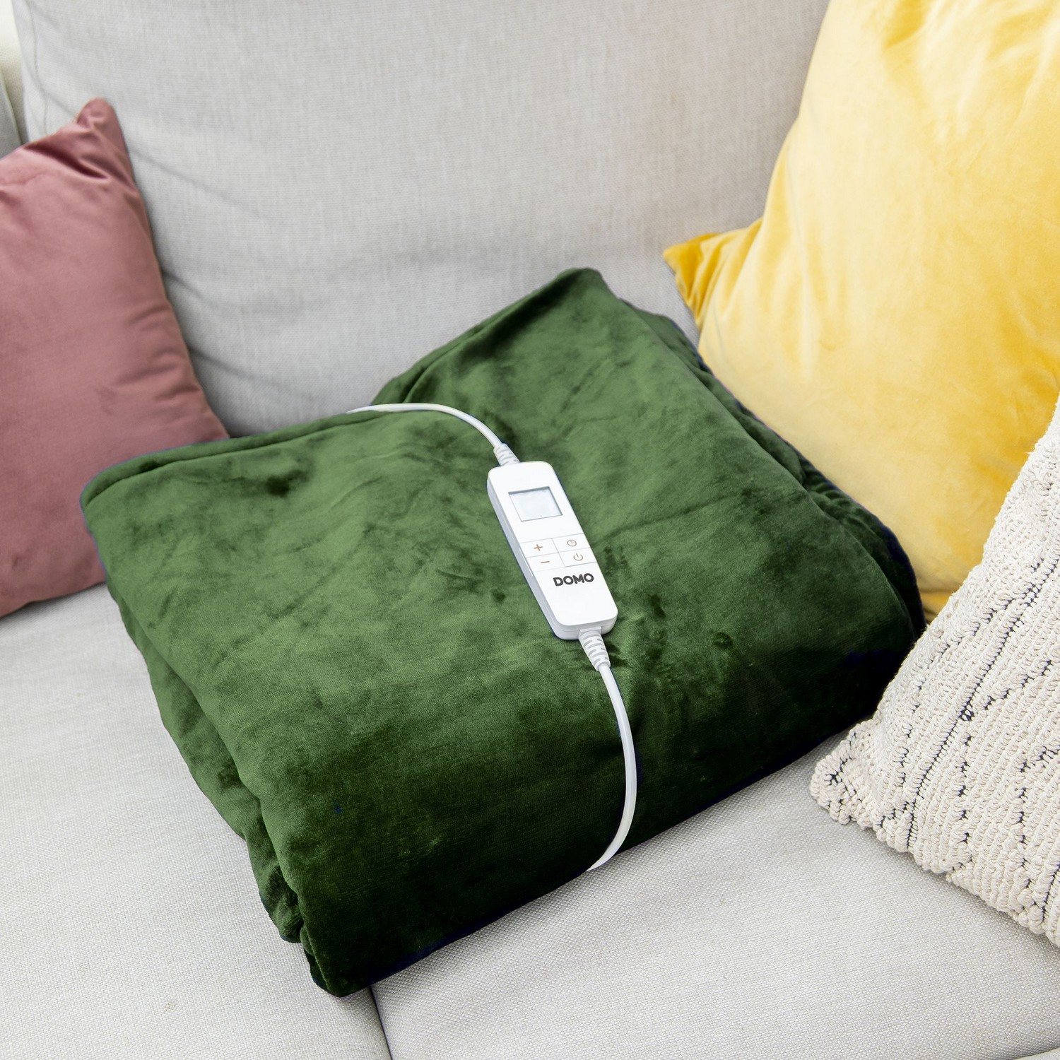 Couverture chauffante verte sur un canapé gris avec des oreillers. Il y a une télécommande sur la couverture.