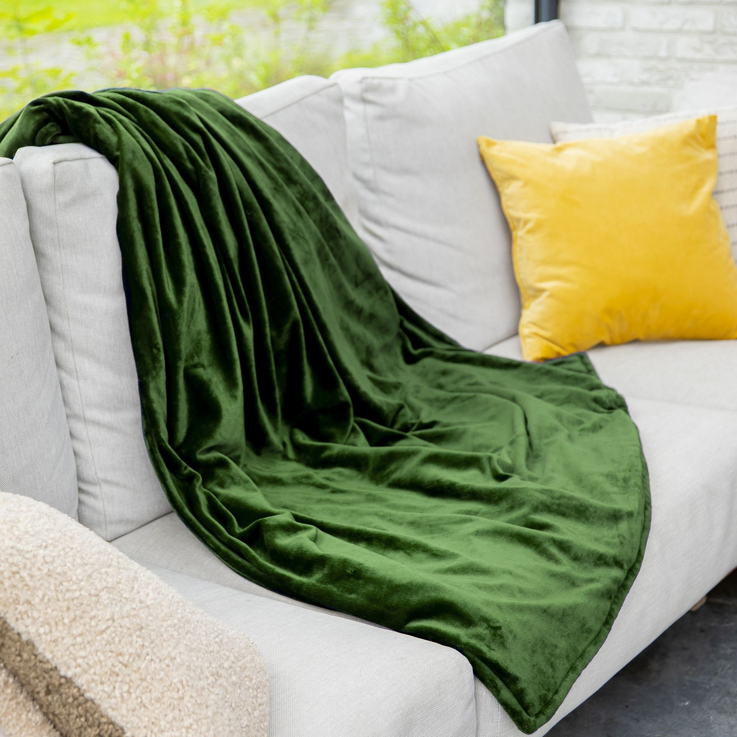 Couverture en velours vert sur canapé blanc, coussin jaune, vue sur fenêtre.