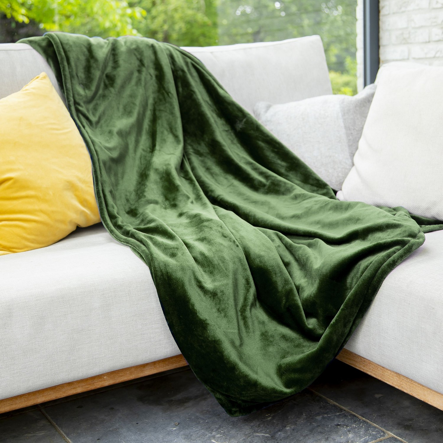 Couverture en velours vert drapée sur un canapé gris clair avec des coussins jaunes et gris.