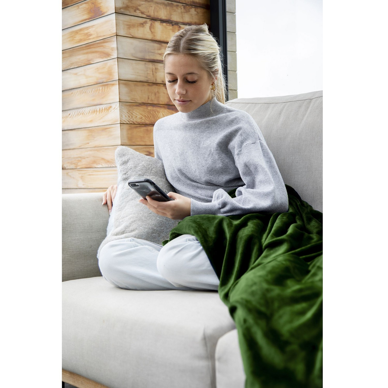 Une femme sur un canapé, regardant son téléphone. Elle est couverte d'une couverture verte et d'un oreiller.