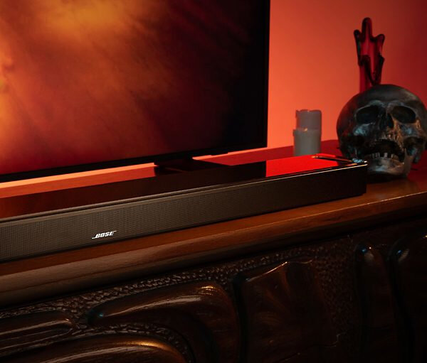 Soundbar na drewnianej powierzchni, telewizor w tle. Czaszka i świece obok. Czerwone oświetlenie otoczenia.