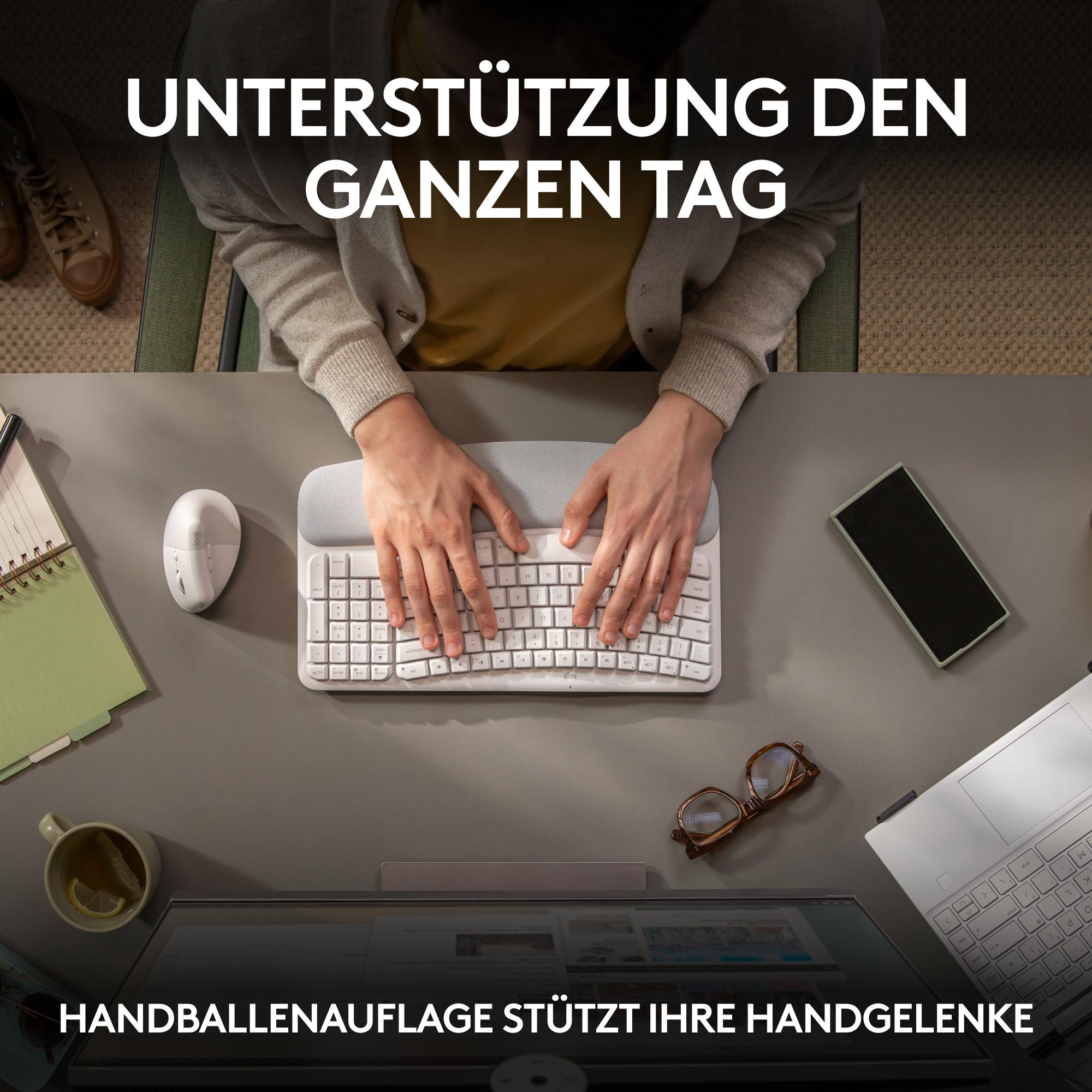 Hände einer Person auf einer Tastatur. Eine Maus, ein Laptop und eine Tasse auf dem grauen Schreibtisch.