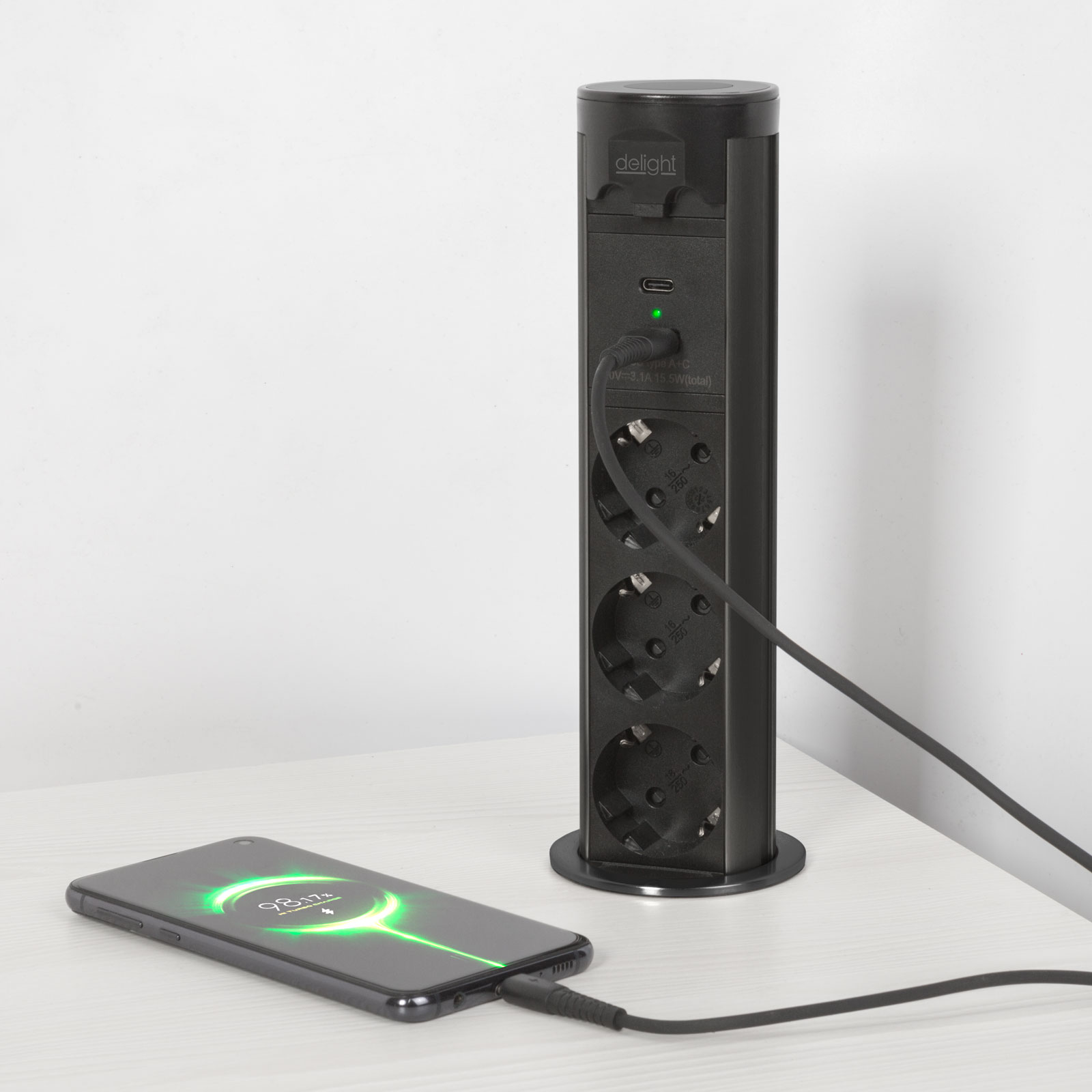 Fekete elosztótorony három aljzattal és USB-porttal. Egy telefon töltődik mellette.