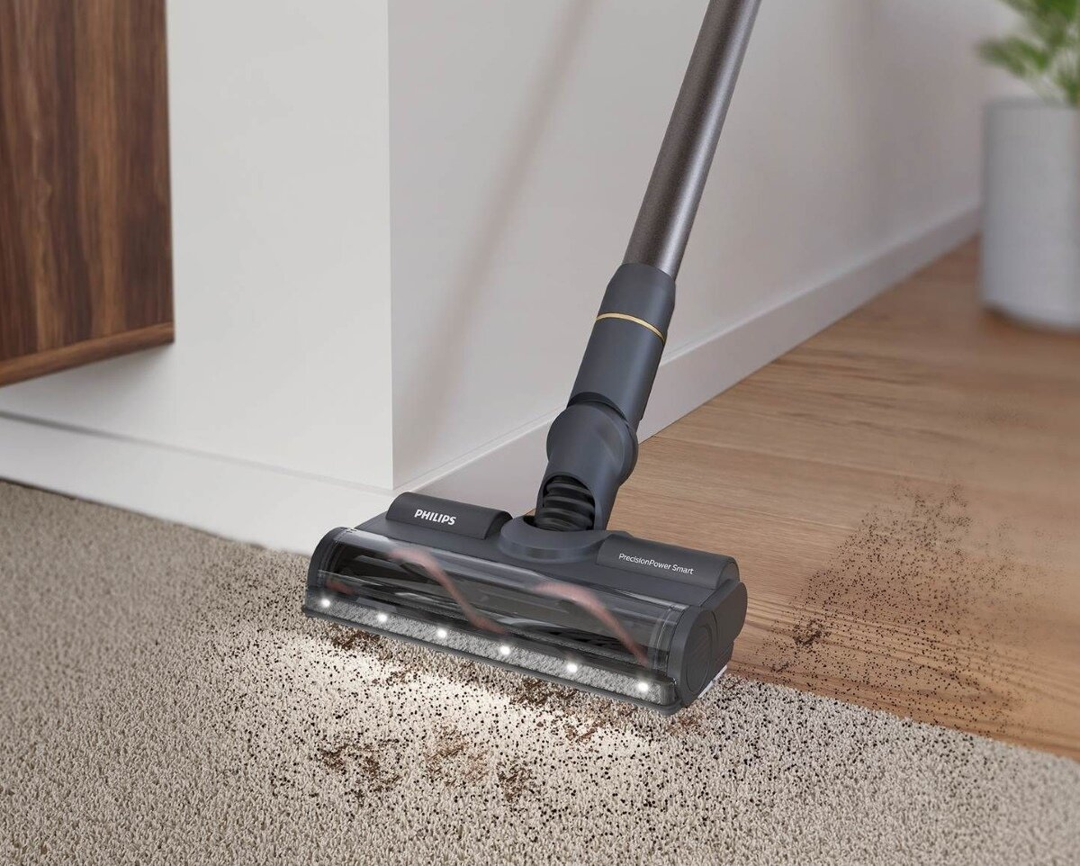 Głowica odkurzacza na dywanie, czyszczenie. Ciemnoszara obudowa, metalowy drążek. Brązowa podłoga i biała ściana w tle.