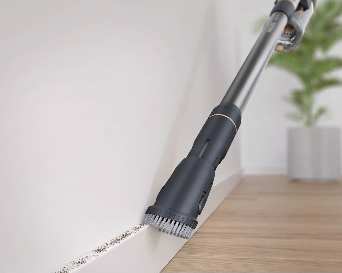 Głowica odkurzacza czyszcząca listwę przypodłogową; małe zanieczyszczenia na podłodze, obok białej ściany i drewnianej podłogi.