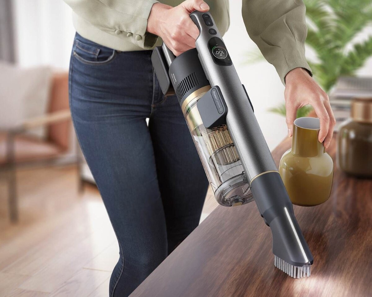 Kobieta trzyma odkurzacz, czyszcząc drewniany stół. W pobliżu jest wazon.