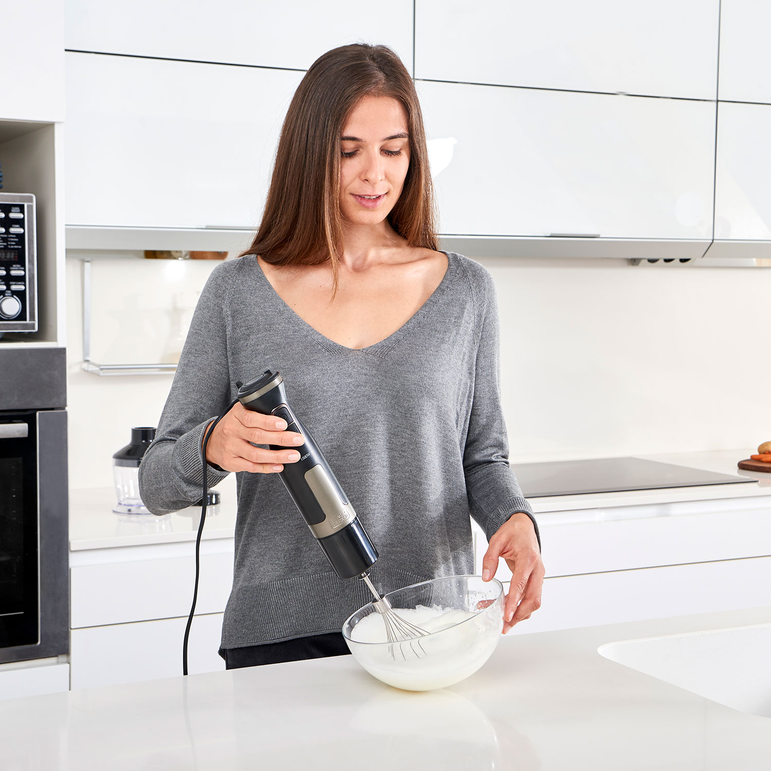 Mujer en suéter gris usando batidora de mano en cocina brillante.