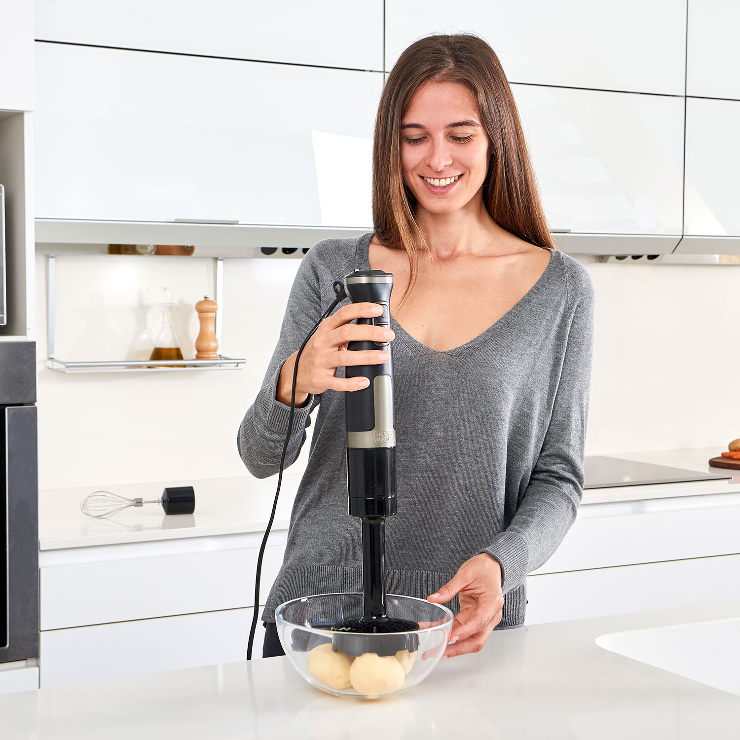 Una mujer usando una batidora de inmersión negra en un bol transparente con papas, sonriendo.