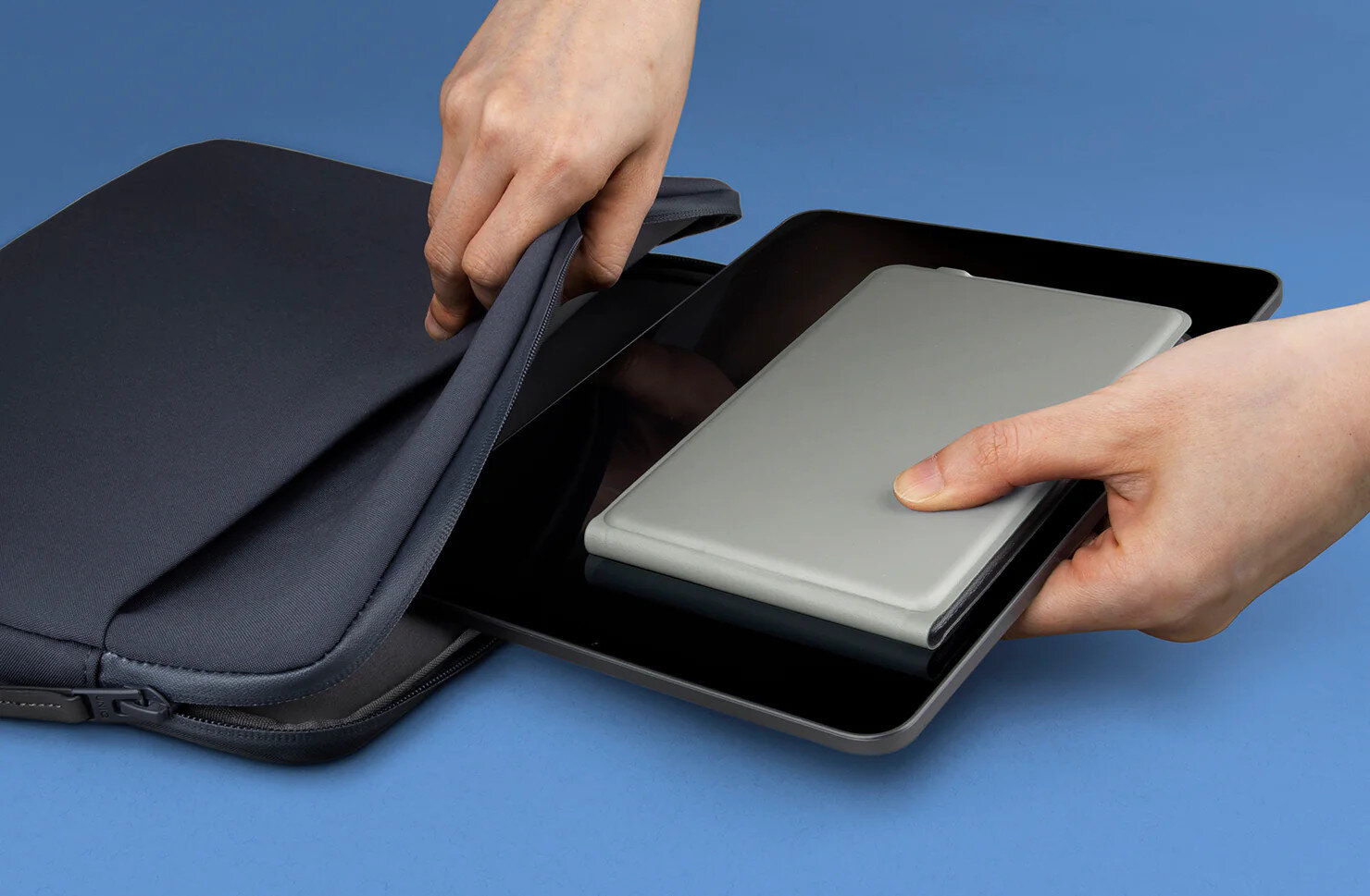Ręce wkładające tablet do ciemnoniebieskiego etui na niebieskim tle.