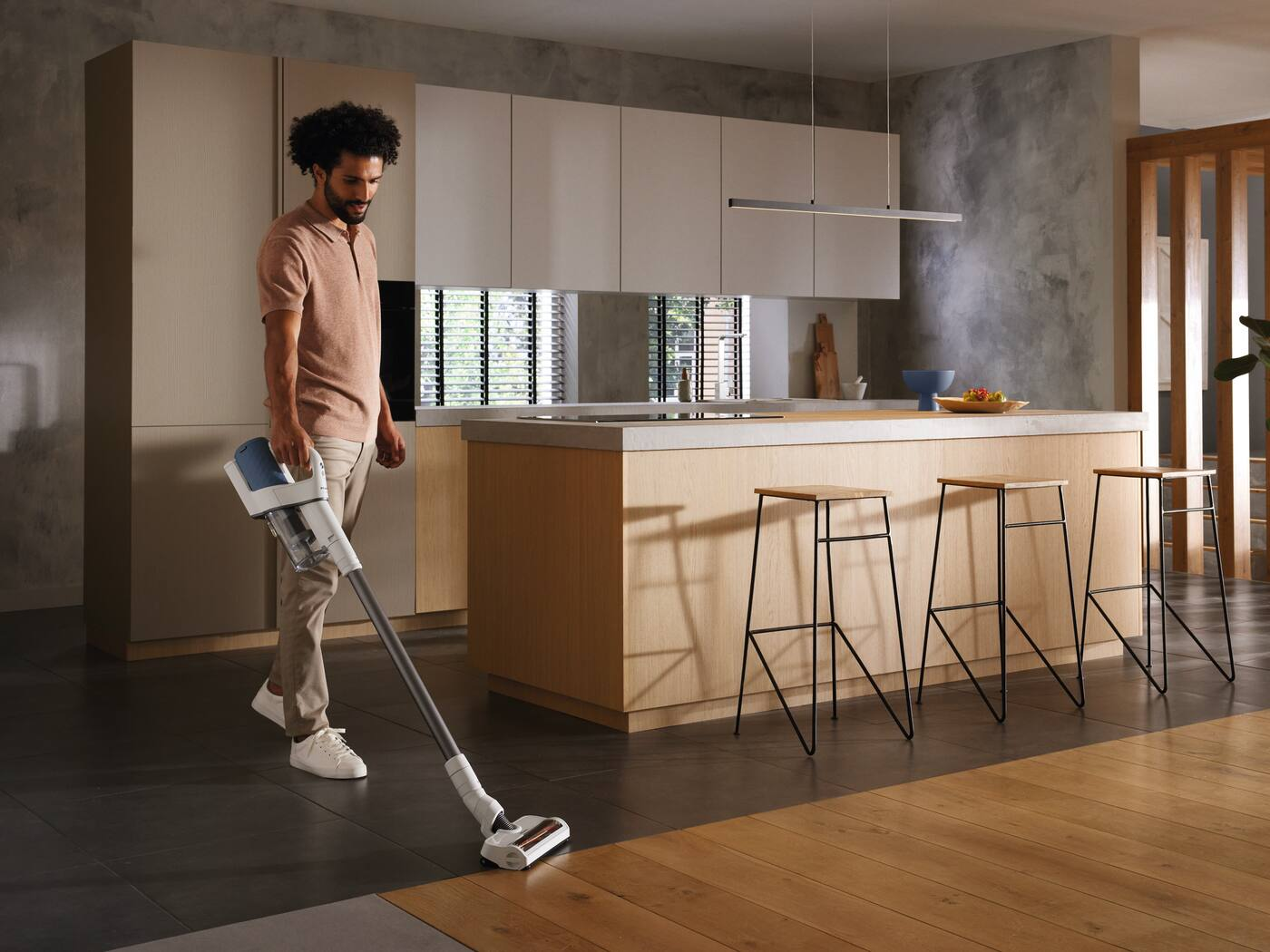 Un homme passe l'aspirateur dans une cuisine moderne, avec un sol en bois clair et des meubles beiges.