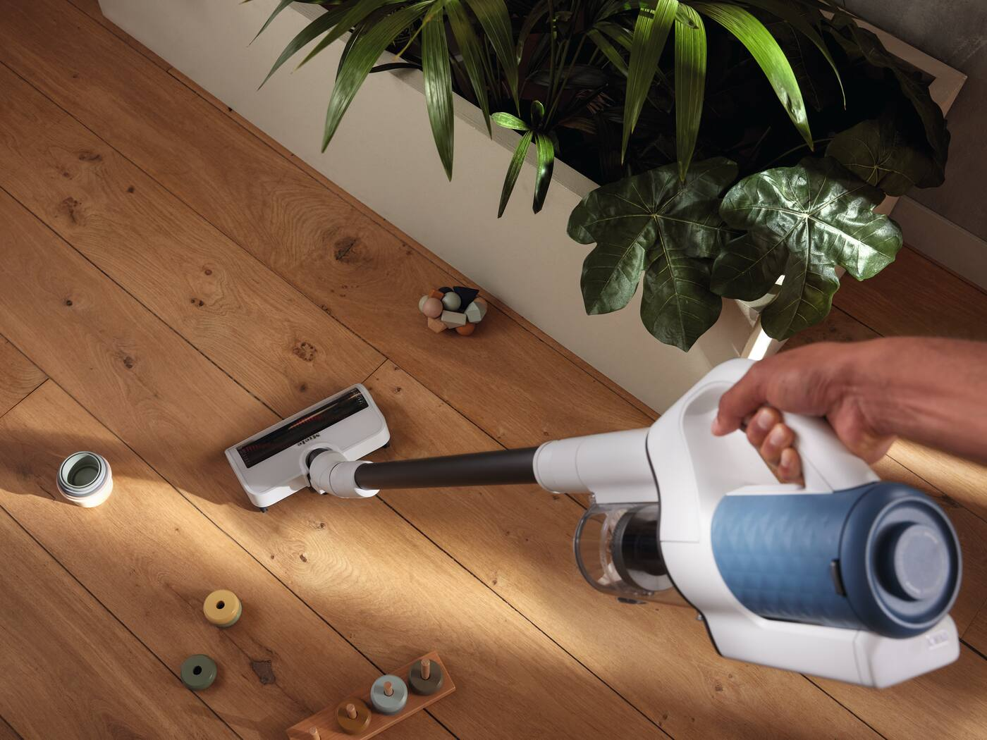 Main utilisant un aspirateur sur un sol en bois. Jouets en bois et plantes vertes en arrière-plan.