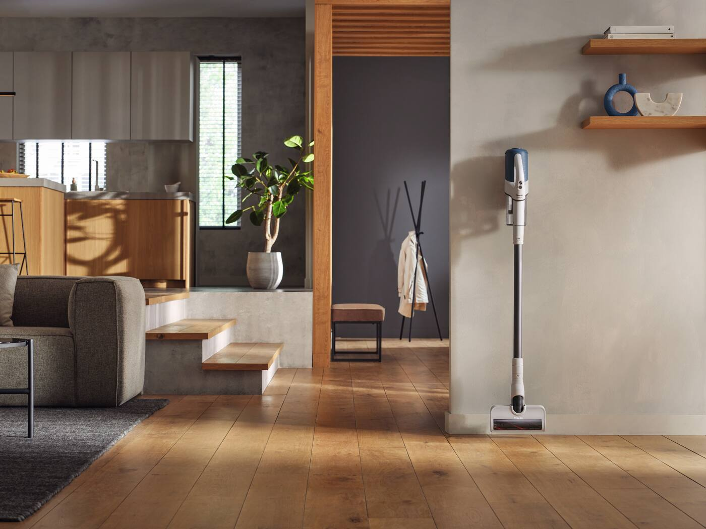 Salon avec parquet. Un aspirateur est appuyé contre un mur. Cuisine en arrière-plan.