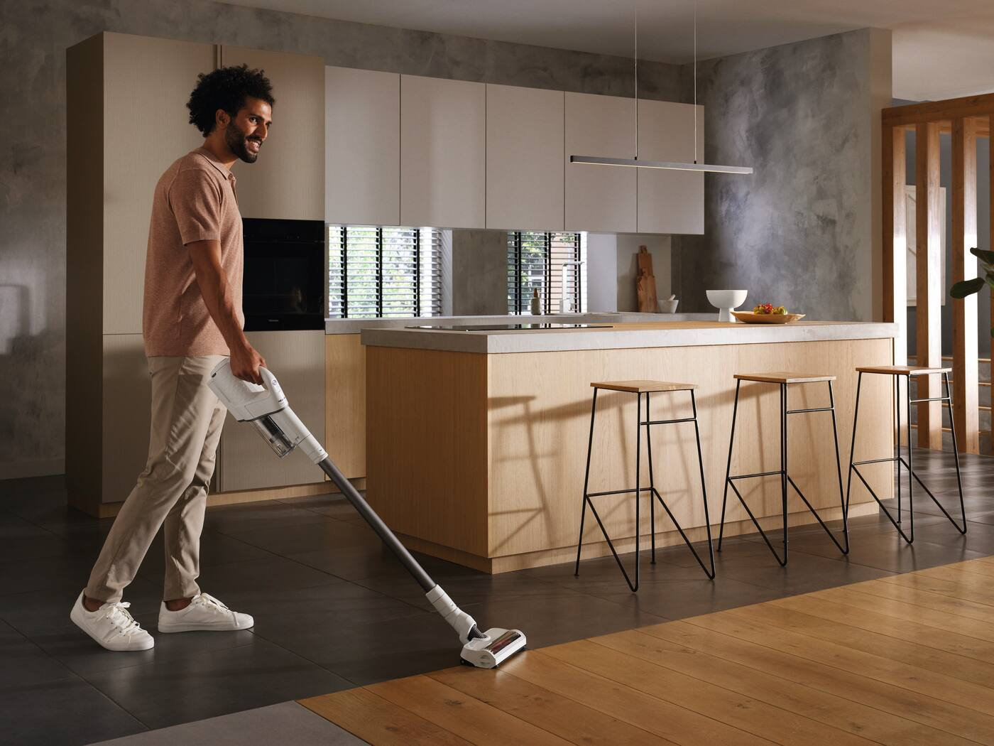 Un uomo aspira il pavimento della cucina. Indossa una camicia rosa e pantaloni color cachi. La cucina ha pavimenti in legno e piastrelle con sgabelli.