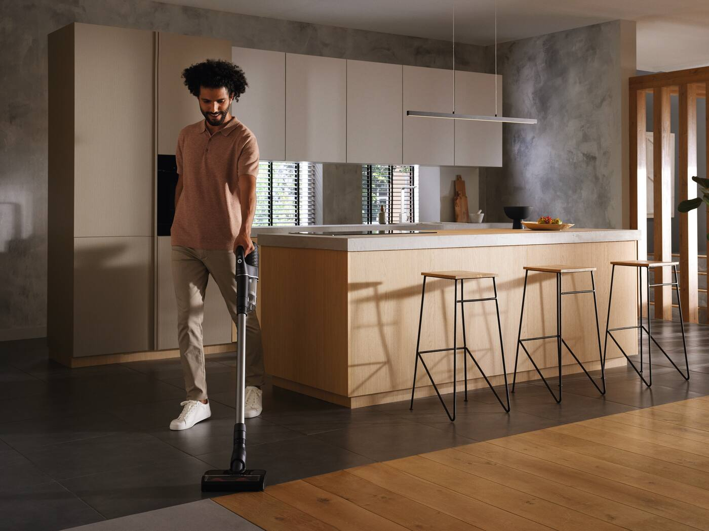 Un uomo in cucina che aspira il pavimento in legno. Ci sono sgabelli e un bancone.