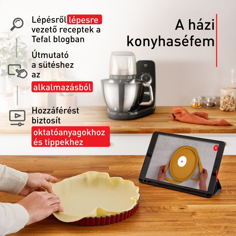 Egy személy tésztát készít egy piros piteformában. Egy táblagép egy főzős videót mutat.