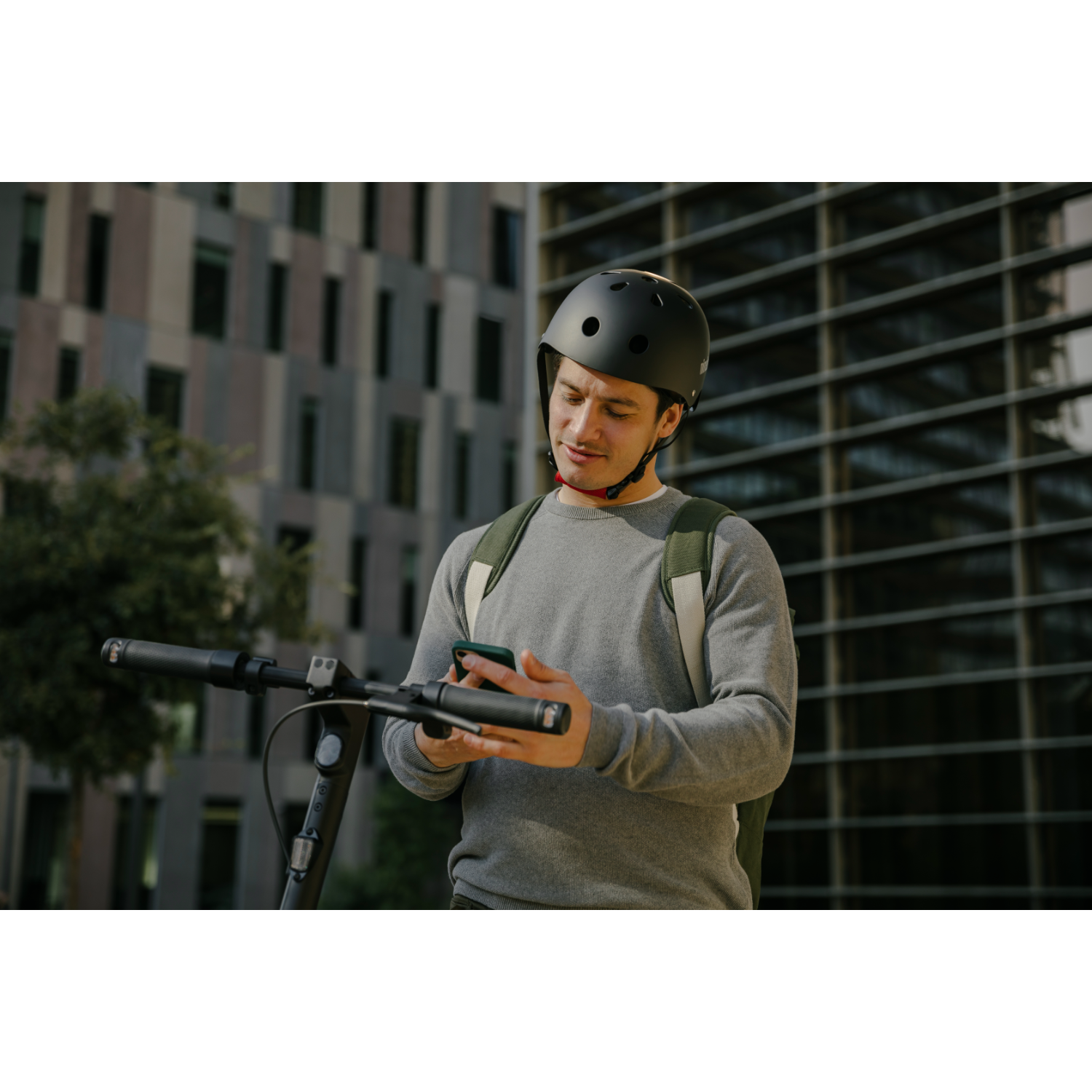 Man met helm op een scooter checkt zijn telefoon. Hoge gebouwen op de achtergrond.