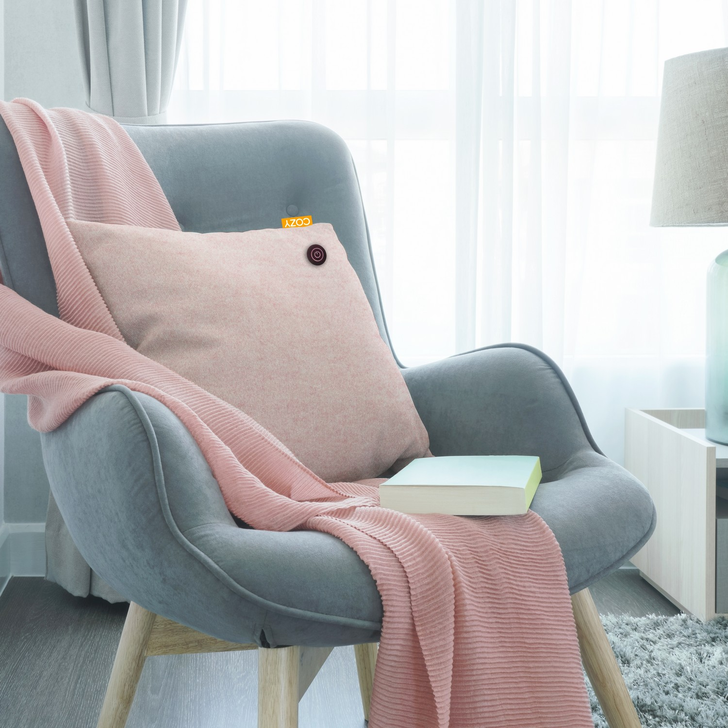 Fauteuil gris avec un coussin et une couverture roses, et un livre. Une lampe à droite.