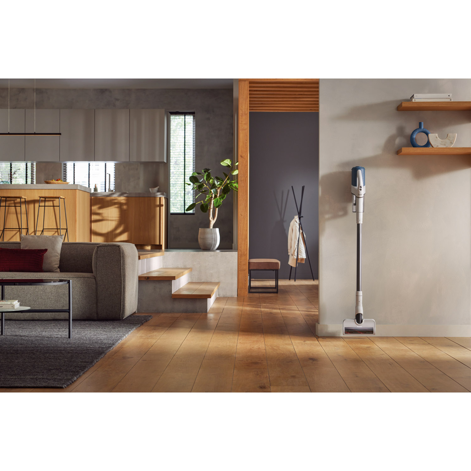 Soggiorno moderno con pavimento in legno, divano grigio, scale per cucina e aspirapolvere.